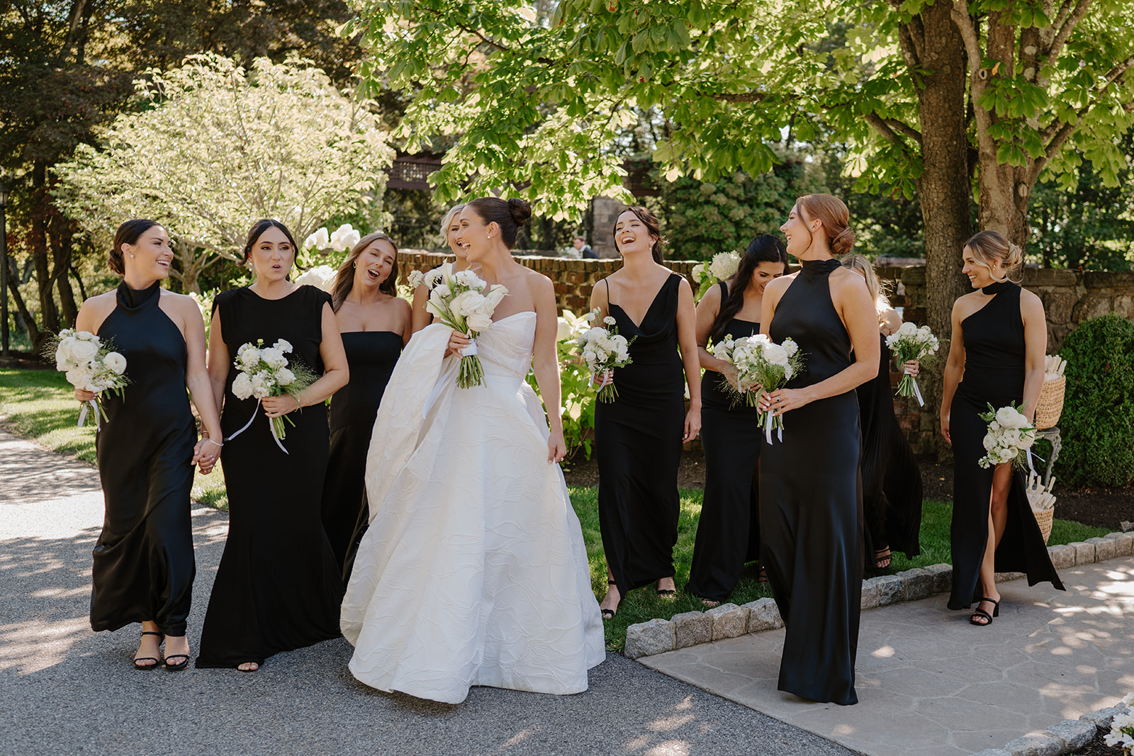 Bride and bridesmaids sharing a candid laugh as they walk together along a paved garden path.